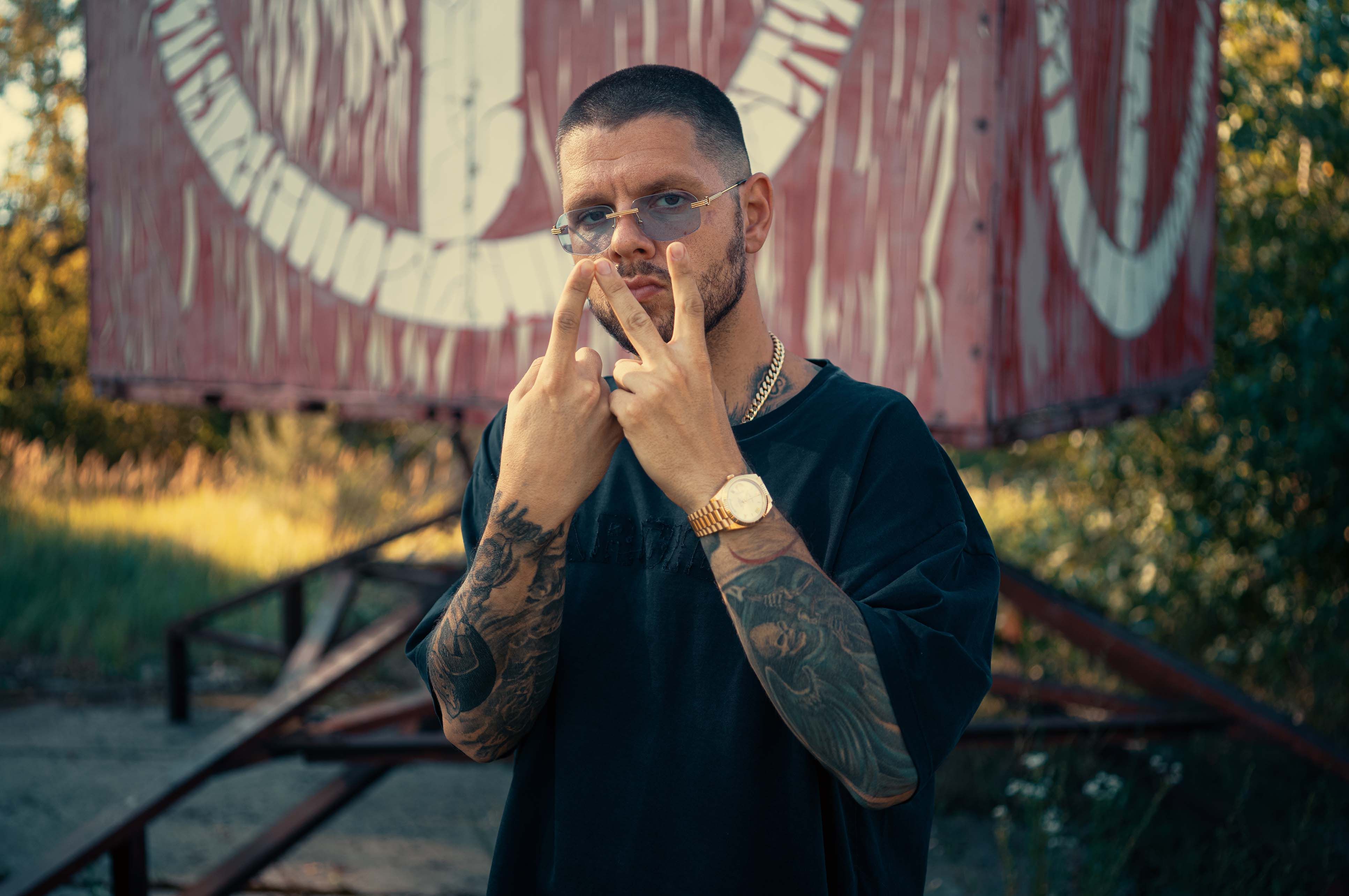 Artysta pozuje na tle drzew. Ma na sobie czarną koszulkę, złoty zegarek i okulary przeciwsłoneczne.