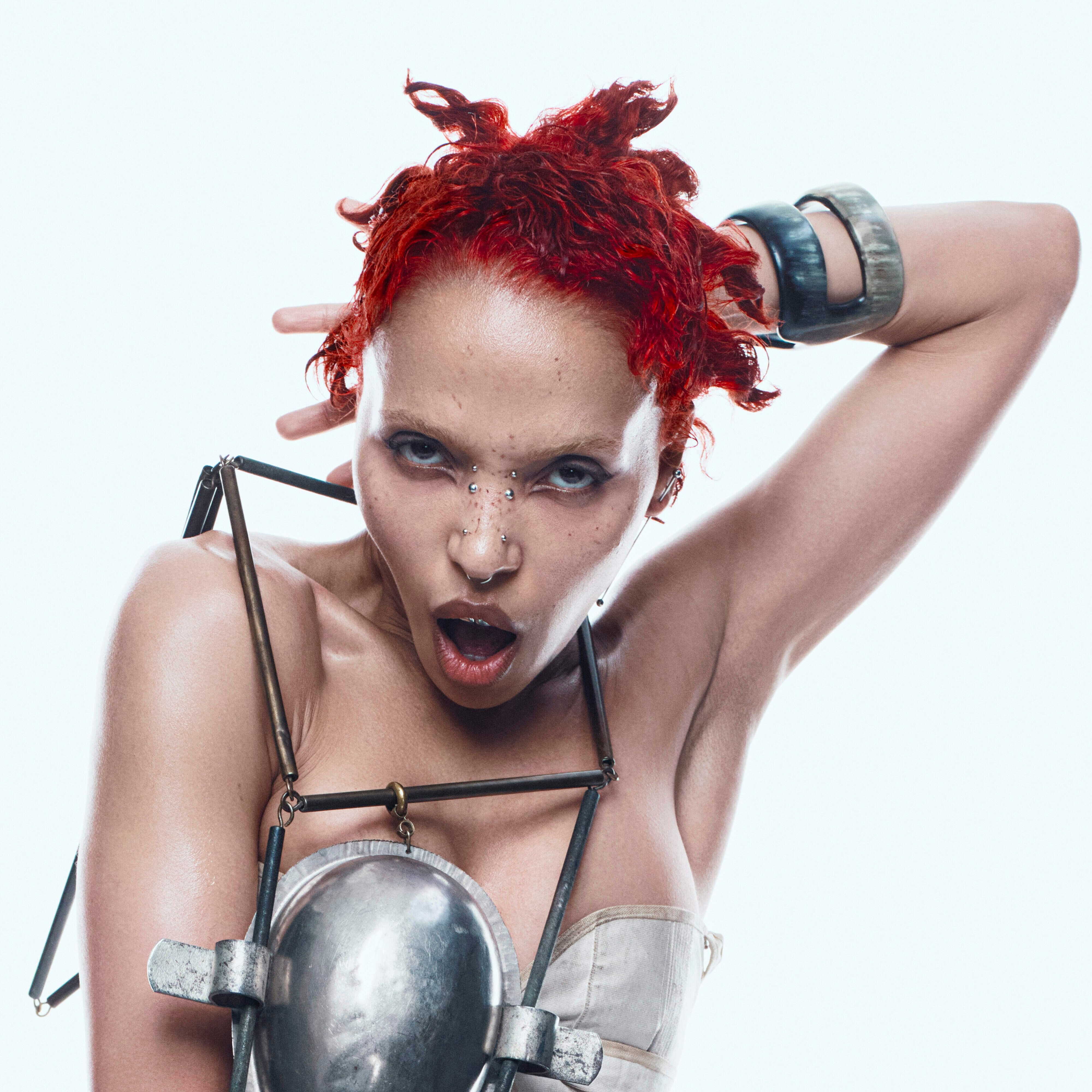 Artystka z zafarbowanych czerwonych włosach i delikatnym makijażu w stroju zrobionym z metalowych elementów.