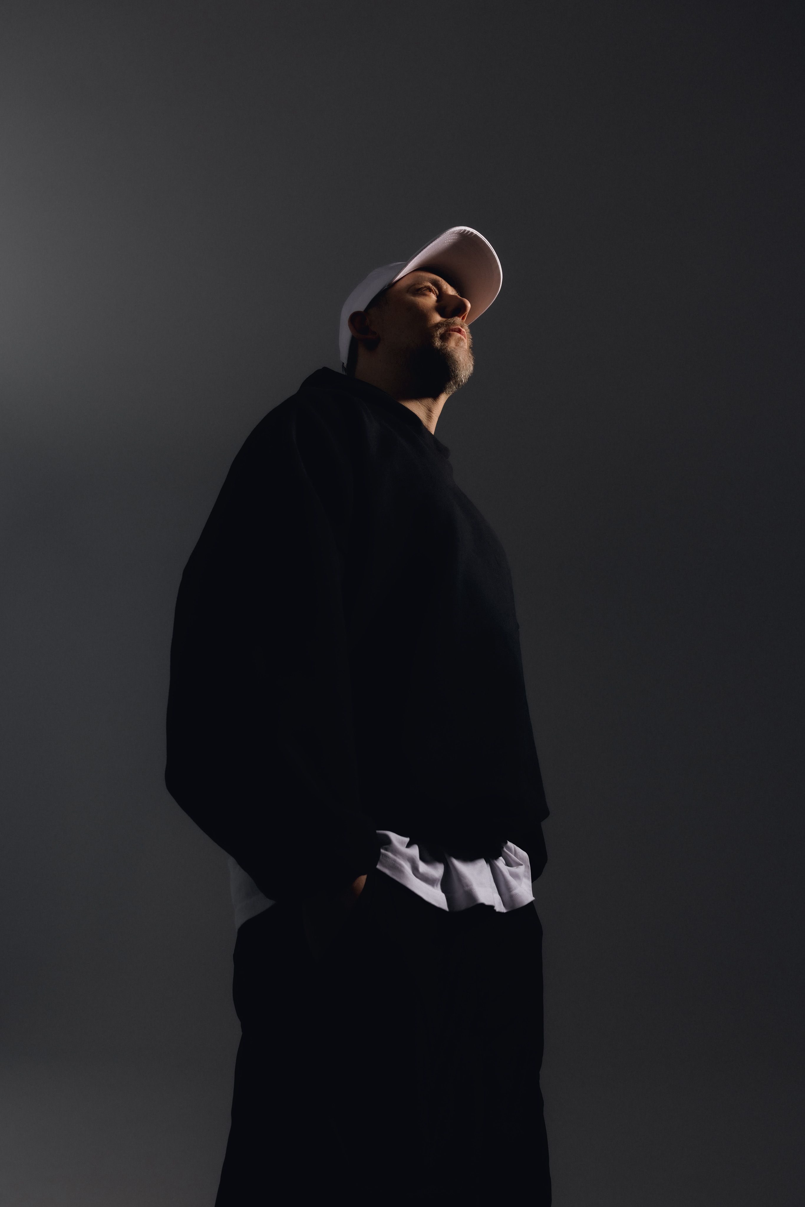 Artysta stoi na czarnym tle, w czarnych ubraniach. Spod bluzy wystaje jedynie błękitna koszula. Na głowie ma różową czapkę.