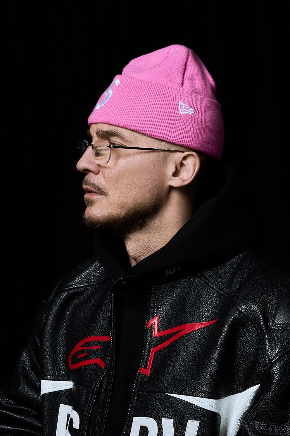 Artysta ustawiony jest swoim lewym profilem do aparartu. Ma na sobie skórzaną czarną kurtkę, różową czapkę i okulary.