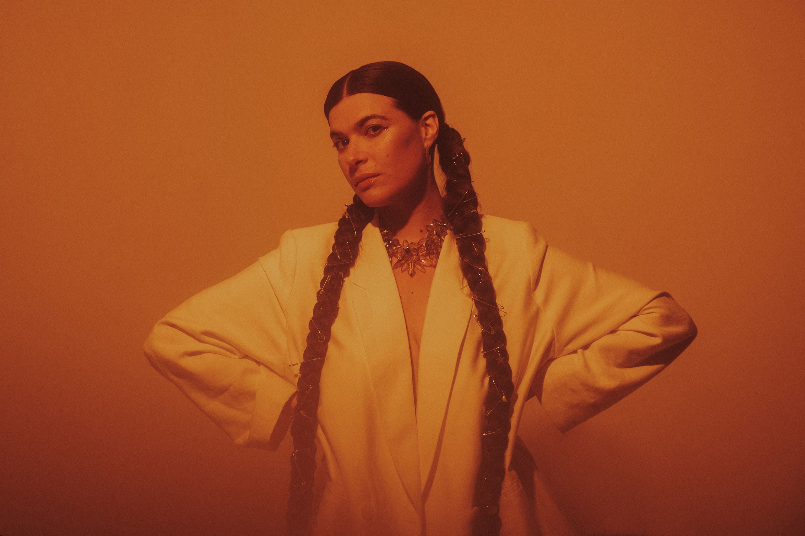 Artystka pozuje na pomarańczowym tle. Ma na sobie długa białą koszulę, opiera swoje ręce na biodrach.