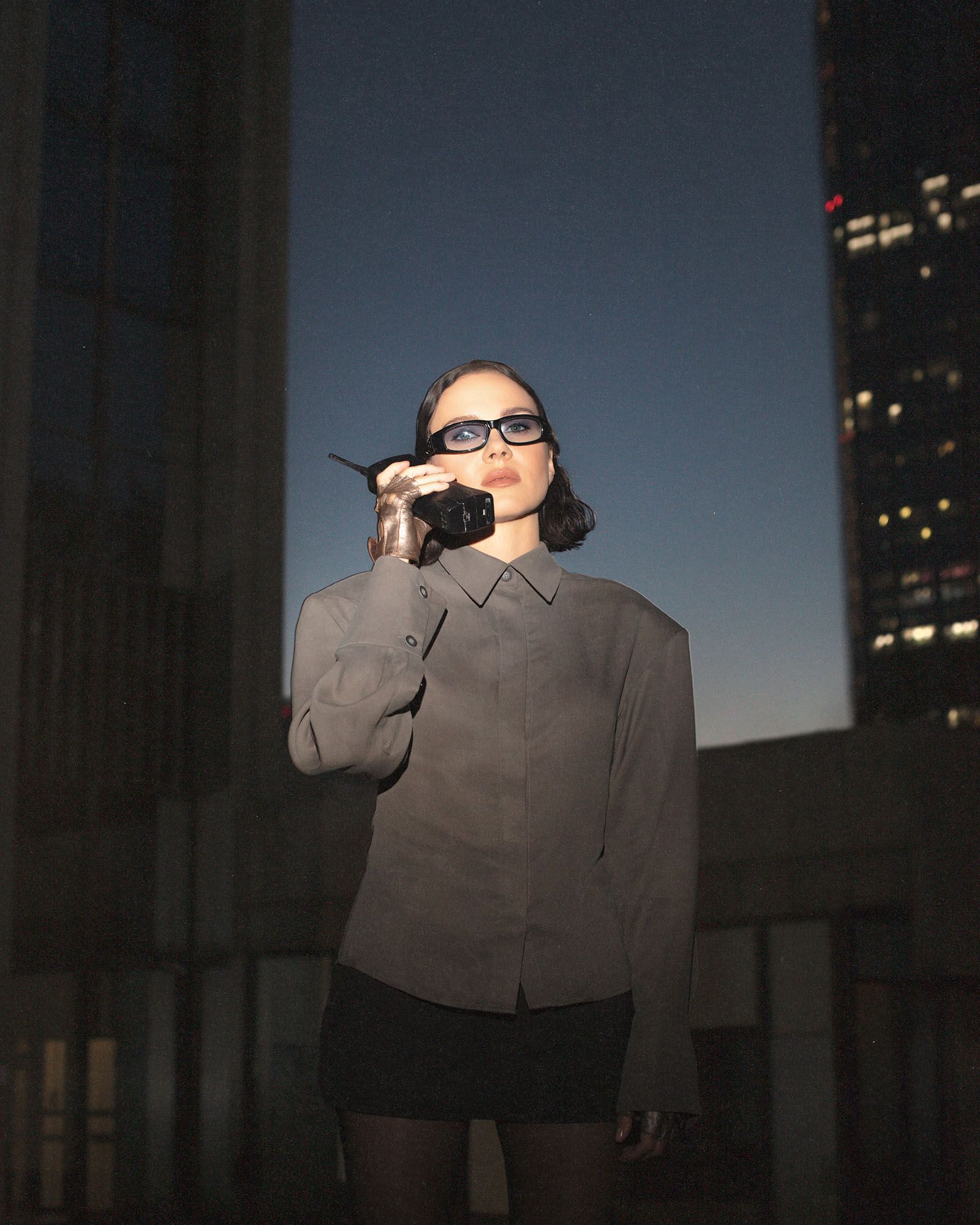 Artyska stoi na tle wierzowców . Ubrana jest w szara koszulę i czarną spódnicę. W ręku trzyma stary telefon.