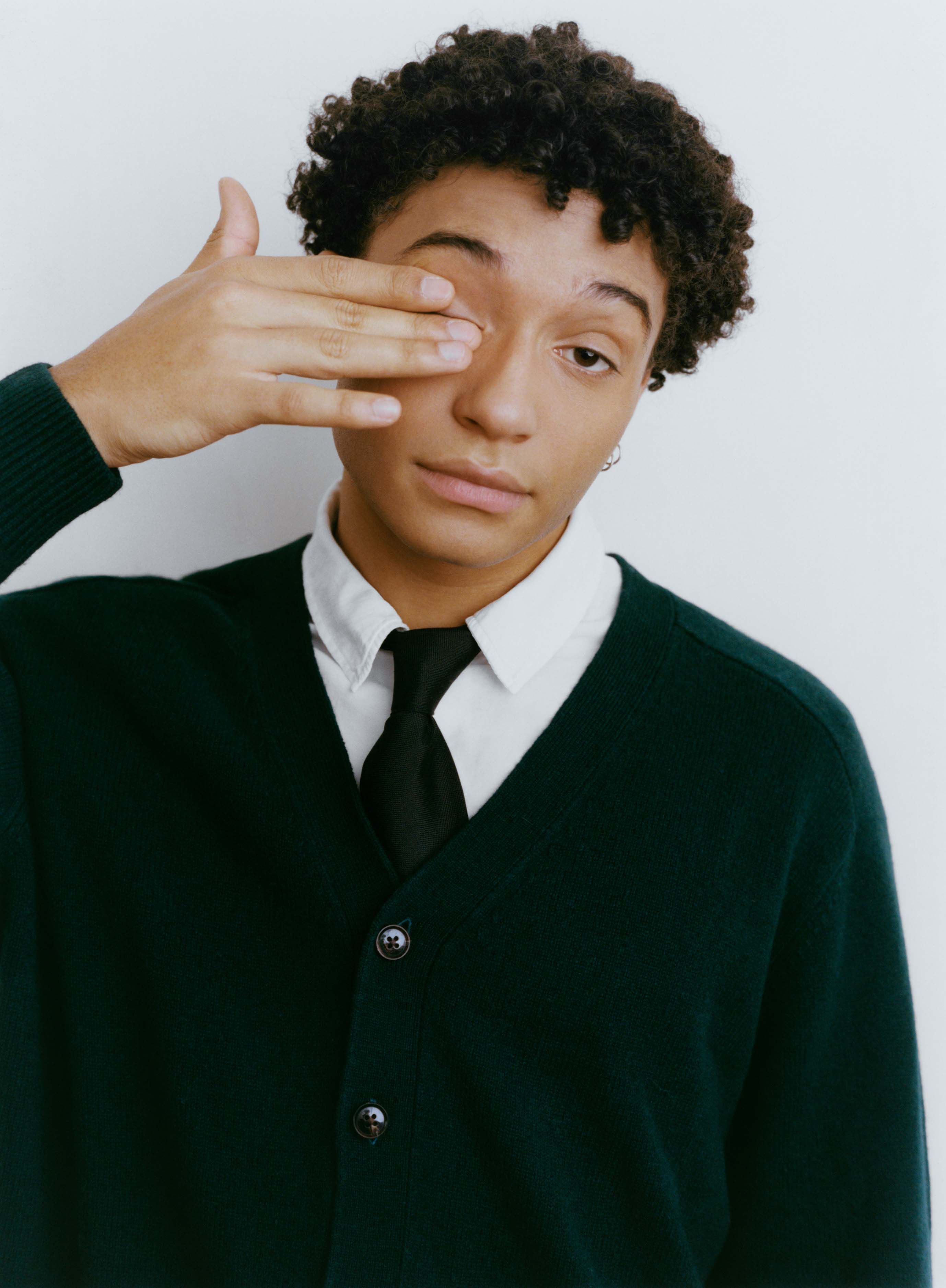 Artysta ubrany na galowo w białą koszulę, czarny krawat i czarny sweter. Prawą ręką załania sobie prawe oko.