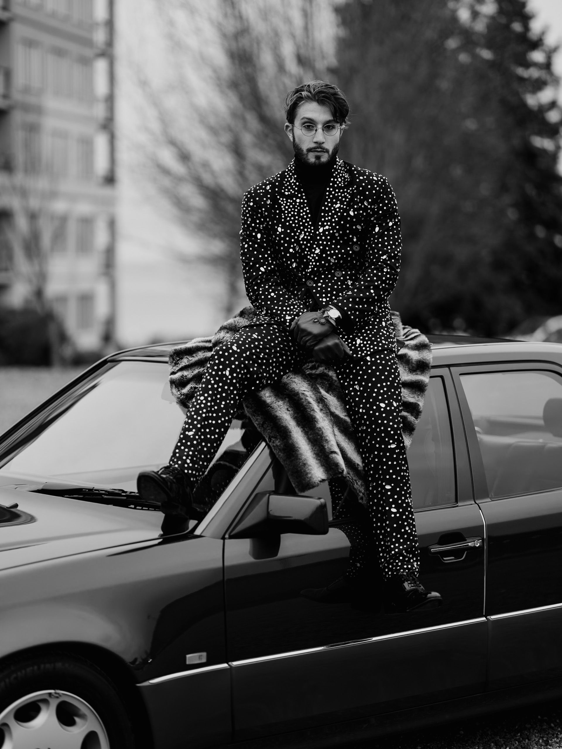 Artystas siedzi na dachu samochodu. Ubrany jest w garnitur pokryty w świecące cekiny.