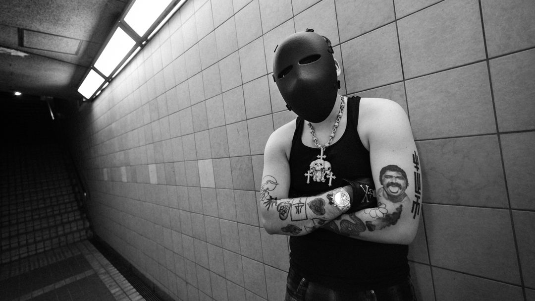 artysta Yeat. Mężczyzna z tatuażami, w czarnej masce, oparty o ścianę