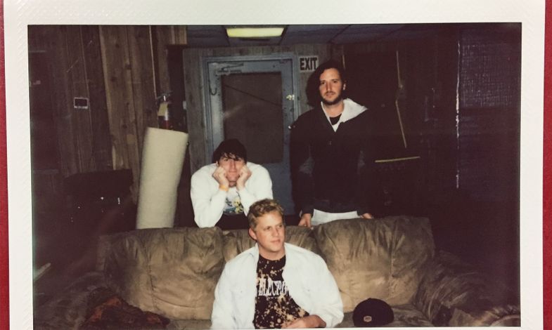 Zdjęcie przedstawia polaroid, w którym trzech artystów siedzi na kanapie vintage