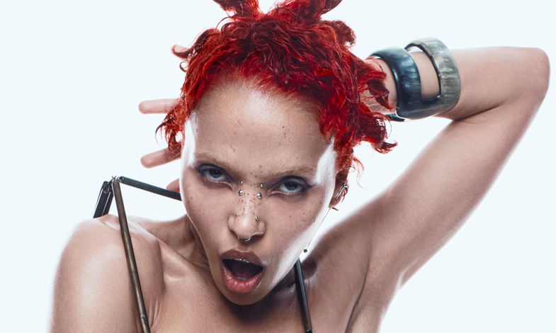 Artystka z zafarbowanych czerwonych włosach i delikatnym makijażu w stroju zrobionym z metalowych elementów.