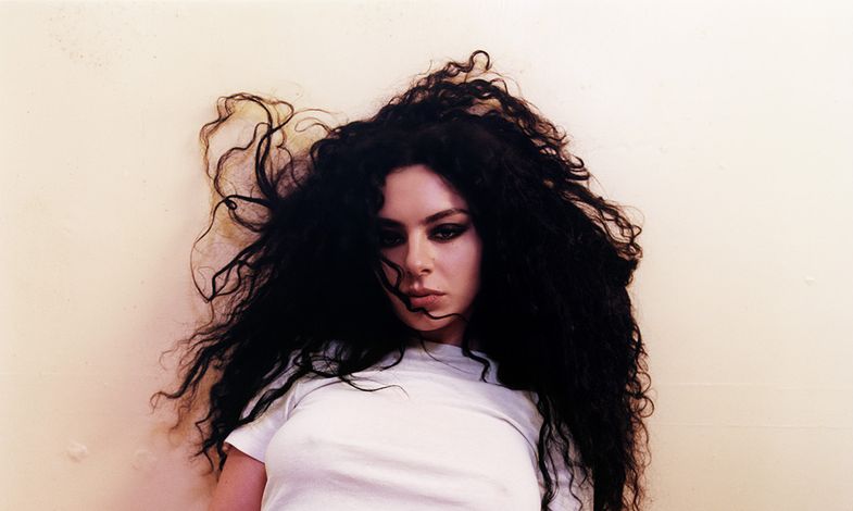 Zdjęcie Charli xcx w długich, czarnych włosach. Artystka jest w białym t-shircie i opiera się o ścianę.