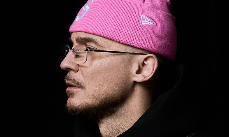 Artysta ustawiony jest swoim lewym profilem do aparartu. Ma na sobie skórzaną czarną kurtkę, różową czapkę i okulary.