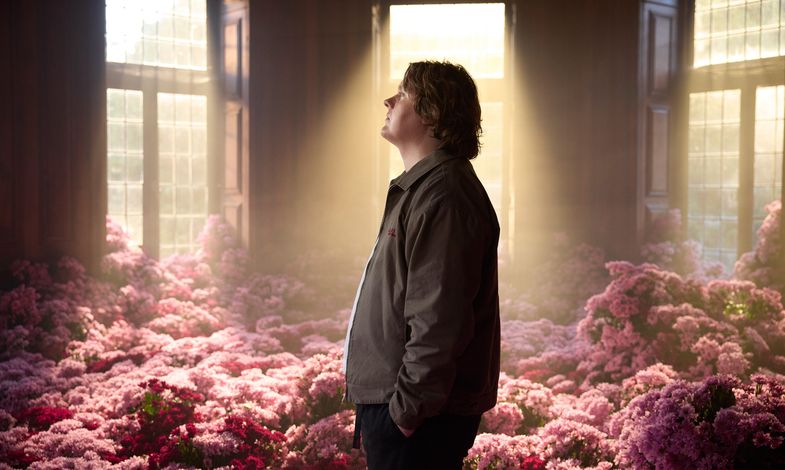 Artysta stoi bokiem w pokoju wypełnionym różowymi kwiatami. Do pomieszczenia wpada słońce przez duże okna.