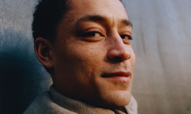 Bliski kadr artysty opierającego się o ścianę. Patrzy prosto w obiektyw z delikatnym uśmiechem. Ubrany jest w zieloną kurtkę.