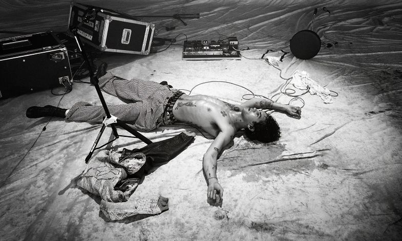 Artysta leży w samych spodniach, bez koszulki na podłodze. Dookoła niego znajdują się instrumenty muzyczne.