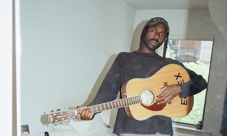 Artysta stoi w sypialni trzymając w ręku gitarę akuratyczną. Za nim znajduje się pościelone łóżko.